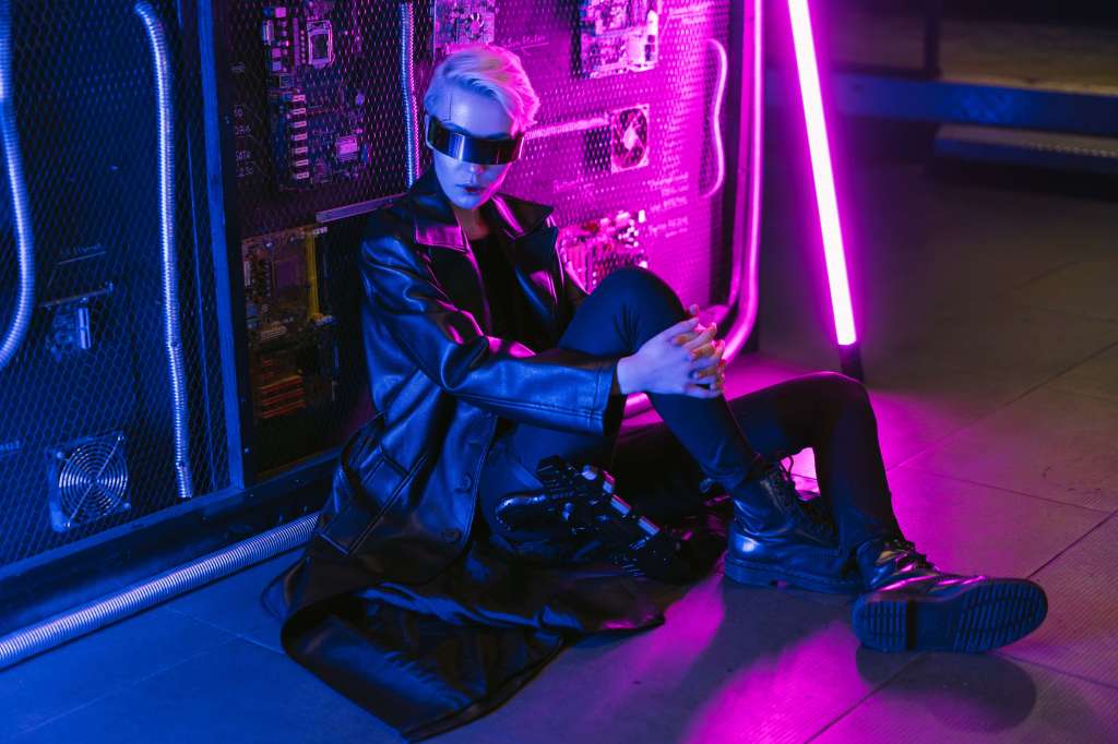 A woman wearing a futuristic visor over her eyes sits on the floor near a pink neon machine.