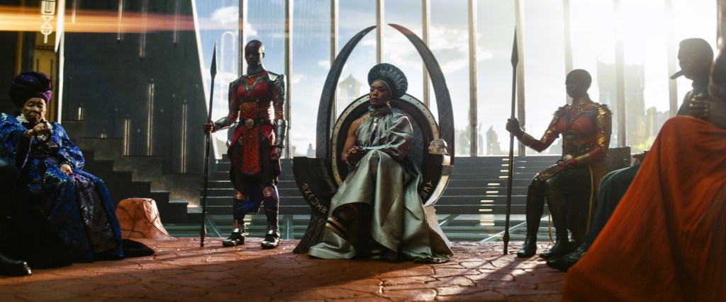 A Black woman in royal dress sits on a throne surrounded by bald female warriors armed with spears.