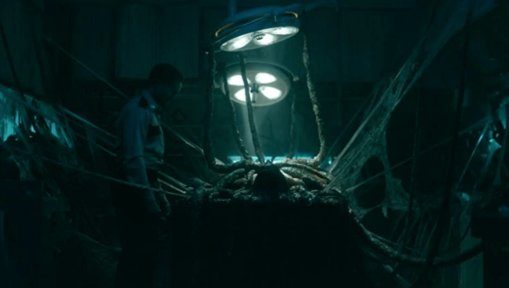 A body sits on a surgical table with large, alien tentacles protruding from the torso and onto every wall.