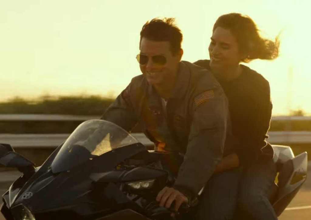 Maverick rides his motorcycle with a beautiful woman on the rear. Their hair flows in the wind as both of them smile.