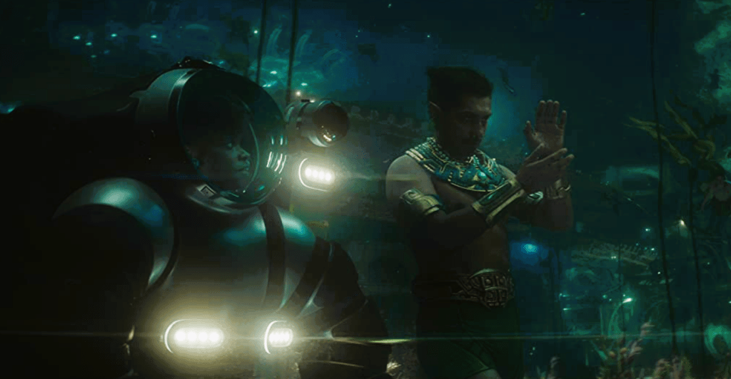 Black woman in deep-sea diver suit standing next to armor clad Latino man underwater.
