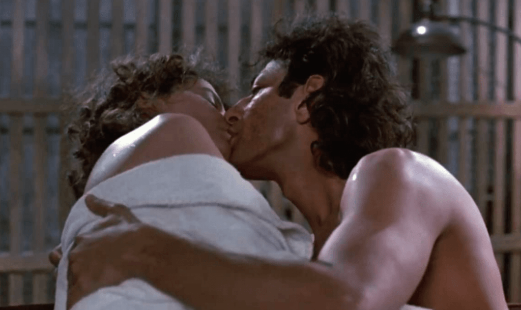 A man and woman kiss passionately inside a studio apartment. Both are half-dressed and covered in a sheet.