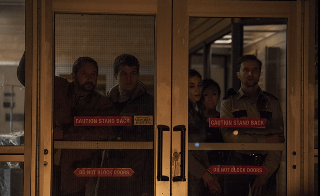 A group of men and women stare outside of sliding doors looking scared.