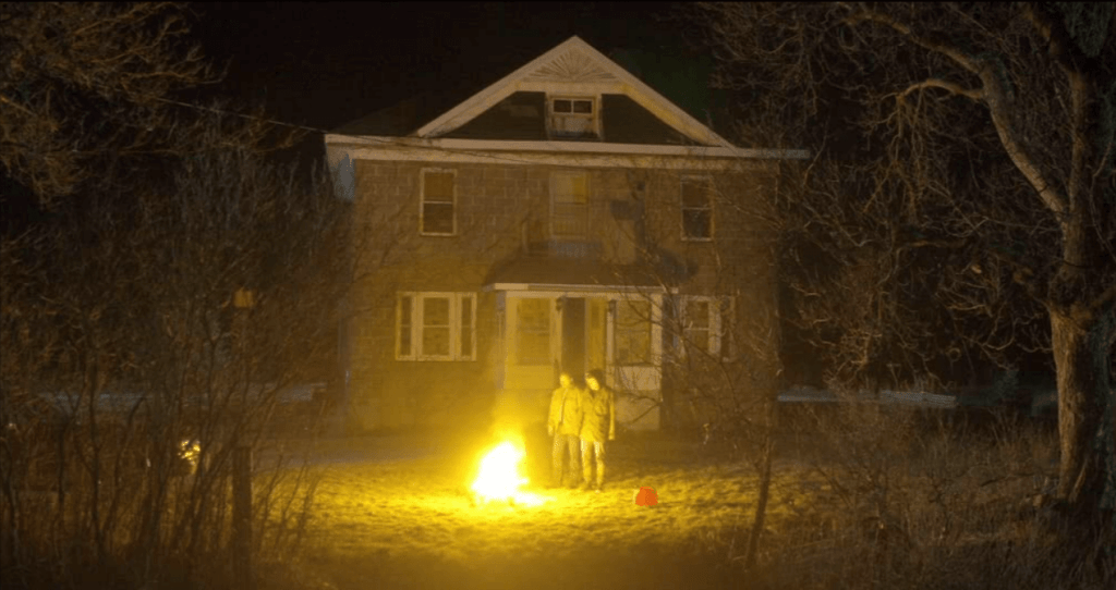 Two men stand outside at night over a burning corpse. 