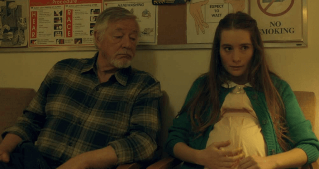 An old man in a flannel shirt sits next to his pregnant granddaughter in a hospital lobby.