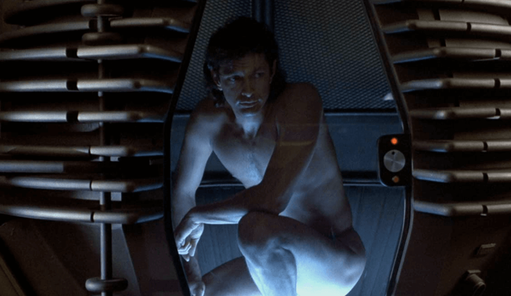 A nude man crouches inside of a pod-shaped teleportation machine with a see-through door.